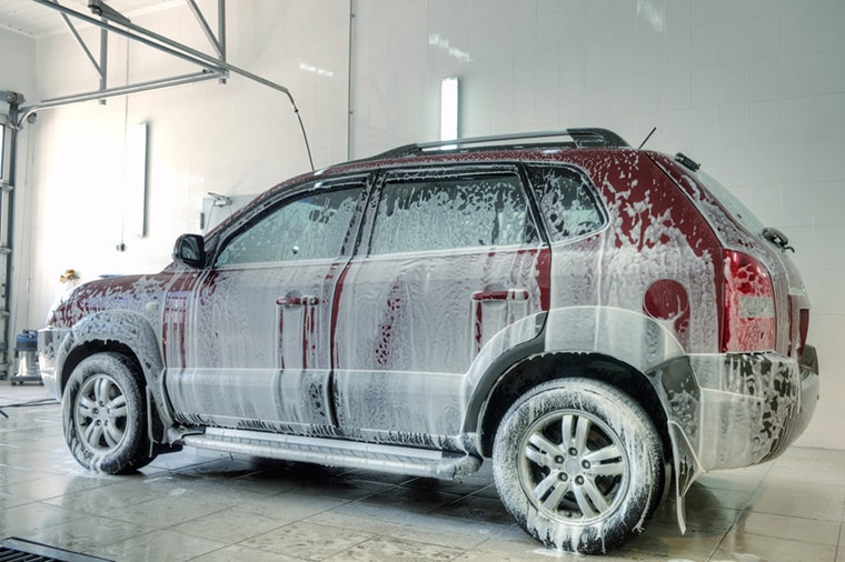 A red SUV is covered in white soap suds while being washed inside a car wash station. The vehicle is on a wet tiled floor, and the background features white tiled walls and industrial lighting.