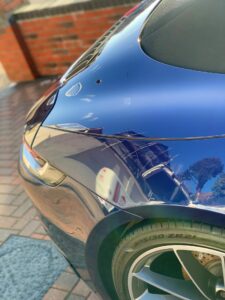 Close-up of the rear section of a shiny blue sports car parked on a brick driveway. The image captures part of the rear fender, tail light, and tire, with clear reflections of a brick wall, a building, and trees on the car’s polished surface.