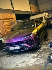 A metallic rainbow-colored Lamborghini SUV is parked inside a garage with a wooden wall on the left and several vehicles, including a caravan, in the background. The garage is dimly lit, and the floor appears to be concrete. The car's license plate reads "RO21E JC.