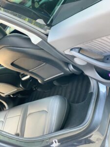 The image shows the interior of a car viewed from an open rear passenger door. The focus is on the back seat, which is upholstered in gray leather, and part of the front seat backrest. The floor is carpeted in dark material with subtle striping.