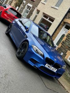 A blue BMW sedan is parked on a residential street, slightly tilted with its front wheels turned outward. Behind it, a red car is also parked on the street. The background includes a row of stone houses with doorways and windows. The road appears wet.