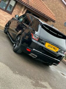 A black Range Rover is parked on a driveway in front of a brick house with a tiled roof. The vehicle has tinted windows and black alloy wheels. The license plate reads "J054 SHU." The driveway and surrounding area are wet, indicating recent rain.