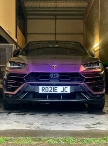 A front view of a purple Lamborghini SUV parked in a dimly lit garage. The license plate reads "RO21E JC". The vehicle's headlights are on, and its aggressive design and sharp lines are clearly visible. The garage has concrete walls and a metal gate on the left.