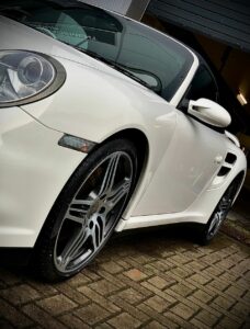 A close-up shot of a sleek, white sports car parked on a brick driveway. The image highlights the car's shiny alloy wheels, aerodynamic design, and part of the front headlight, exuding a sense of luxury and high performance.