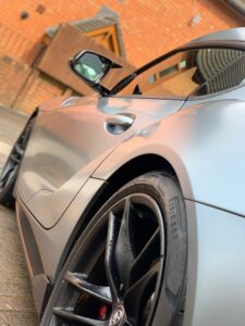 A close-up, angled shot of a sleek, metallic grey sports car showcasing the car's side, rear wheel, and part of the side mirror against a brick building and an angular, wooden structure in the background. The car's tire is marked with a Pirelli logo.