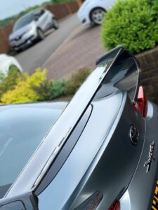 Close-up of the rear spoiler of a sporty car with a sleek design, parked in a driveway. The spoiler is carbon fiber, enhancing the vehicle's aerodynamic appearance. In the background, there are other parked cars and green shrubbery.