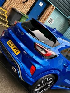 A vibrant blue Ford Puma with a license plate "EG22 RTX" is parked in front of a blue door and a brick wall. A small green garbage bin is visible next to the door, which has a sign reading "Nelson Projects." The car's rear is prominently displayed.