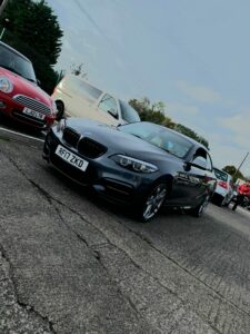 A dark grey BMW parked on a street in front of a white van, a red car, and a red Mini with a visible license plate. The sky is partly cloudy and there are trees in the background.