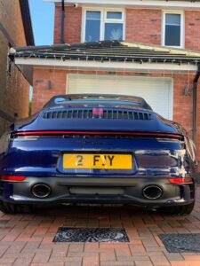 A sleek, dark blue Porsche sports car is parked in front of a modern brick house with white garage doors and holiday lights hanging from the roof. The license plate reads "2 FJY." The car is positioned on a brick-paved driveway.