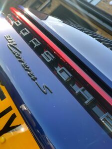 Close-up of the rear end of a dark blue Porsche 911 Carrera S, showcasing the car's emblem and model name. The image captures the sleek, glossy finish of the vehicle, with part of the yellow license plate visible at the bottom.