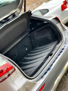 Open trunk of a silver car with a clean, empty, and spacious cargo area. The trunk door is raised, revealing the interior of the trunk lined with a dark material. Another white car is parked nearby, visible in the background.