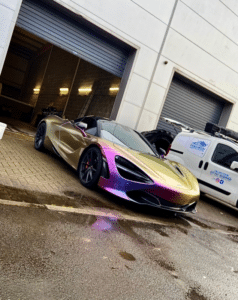 A sleek, iridescent sports car with shades of gold, purple, and green is parked in front of a building with metal roll-up doors. A white van with a partial logo on its side is parked nearby, and the ground is wet, suggesting recent rain.