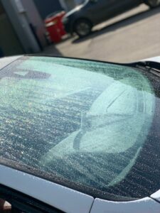 Close-up of a car windshield covered in water droplets. The sun is shining, creating a slight glare and reflections on the glass. A few vehicles and a red object are blurred in the background.