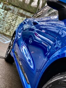 A vibrant blue car is parked on a wet street. The car's surface reflects nearby vehicles and trees, creating a mirrored effect. The background features a stone wall and a metal fence with leafless trees visible above. The scene has a glossy and reflective quality.