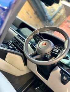 Interior view of a car featuring a leather-wrapped Porsche steering wheel with the emblem prominently displayed. The dashboard and console are modern with sleek lines and details, including an electronic display and control buttons, set against a light-colored upholstery.