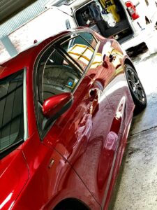 A red car is parked in a garage, reflecting the surroundings on its shiny surface. In the background, the back of a white van with its doors open shows various tools and equipment inside. The environment appears to be a vehicle maintenance or detailing area.