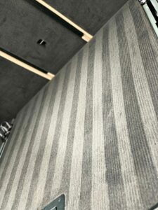 An image of a carpeted floor with a striped pattern in varying shades of gray. There are a few metallic fixtures and the base of some furniture visible at the corners. The image is taken from a close-up perspective.