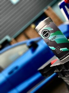 A canister labeled "Synergy" rests on a surface with a design featuring shades of blue and green. In the background, a blue car is parked, slightly out of focus, against a building with gray siding and a brown brick base. The scene has a slightly tilted perspective.