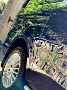 Close-up of the side of a shiny, dark-colored car with reflections of green trees and a wooden fence on the surface. The focus is on the wheel and part of the car's body, highlighting the clean, polished finish.