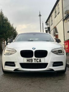 A white BMW is parked on a residential street with trees and houses on either side. The car's front license plate reads "H19 TCA." The sky above is overcast.