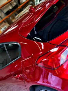 A shiny red car is parked inside a garage. The focus is on the rear section, highlighting the sleek design, red taillights, and reflective surface. Shelves with various items are visible in the background.