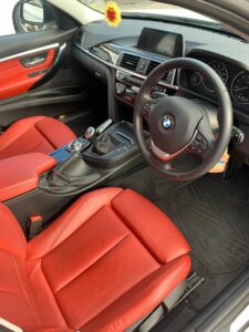 Interior of a BMW car with red leather seats and black trim. The steering wheel has the BMW logo, and the dashboard features various controls and a digital display screen. A flower-shaped air freshener hangs from the rearview mirror. The gear shift is visible.