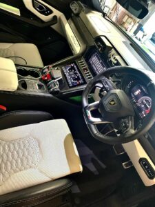 Interior view of a luxury car, featuring a multi-function steering wheel, a digital dashboard, and a central console with various controls and displays. The seats are upholstered in a patterned material with a combination of light and dark colors.