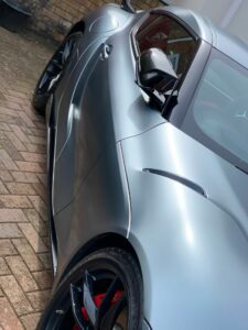 Close-up of a sleek, silver sports car parked on a brick driveway. The car's side view mirror, aerodynamic curves, and black alloy wheels with red brake calipers are prominently featured. The shiny finish and modern design elements convey a sense of luxury and performance.