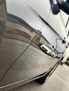 A reflective side panel of a car mirrors nearby vehicles and houses along a street. The close-up perspective captures the smooth, polished surface of the car, providing a distorted but clear view of the surroundings.