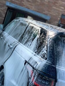 A black SUV is partially covered in soap suds, indicating it is being washed. The vehicle is parked in front of a brick building with a green door and window. Sunlight reflects off the wet surface of the car, producing a shiny effect.