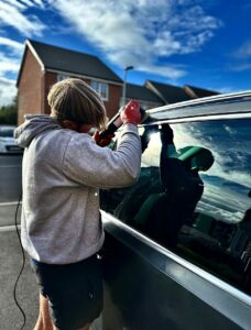 A person wearing a grey hoodie and red gloves installs a black window on a car using a tool. The scene appears to be outdoors near residential buildings. The sky is partly cloudy with sunlight creating reflections on the car's surface.