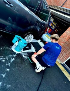 A person wearing a blue shirt and black shorts is washing a car in a driveway. They are using a hose and a blue bucket filled with soapy water. The garage door is open, revealing various items inside. Soap suds are visible on the ground.