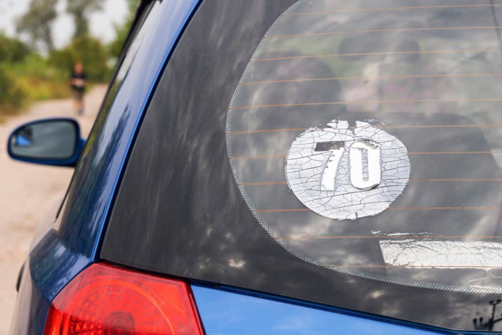 Close-up of a blue car's rear window featuring a cracked circular sticker with the number "70" on it. The car is parked on a dirt road with trees in the background.