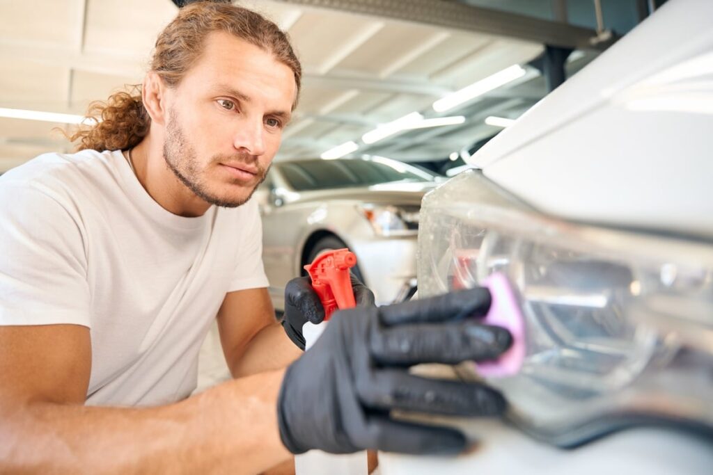 A man with long hair and a white t-shirt carefully cleans a car's headlight with a pink cloth. He wears black gloves and holds a spray bottle, focusing intently on his work in a well-lit garage.