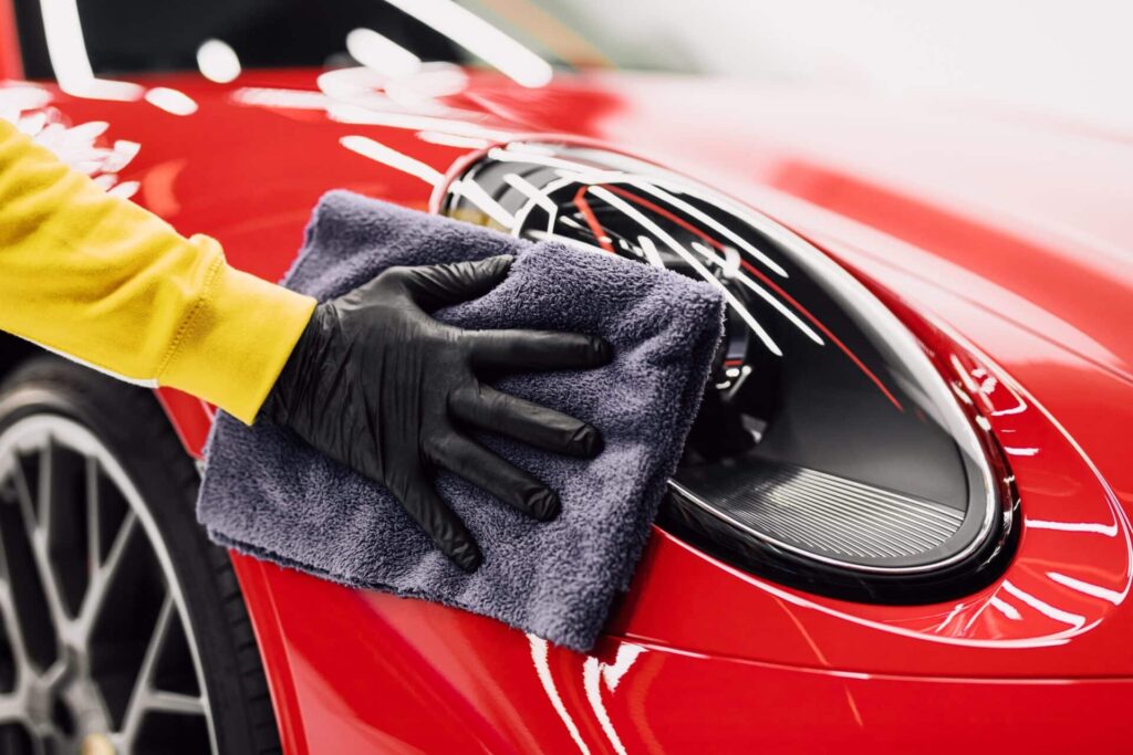 A person wearing a yellow sleeve and black glove polishes the headlight of a red sports car with a gray cloth. The car's sleek, glossy finish reflects the light.