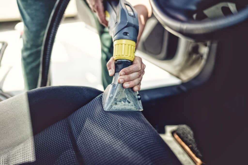 Close-up of a person using a vacuum cleaner to clean a car seat, protecting your car's upholstery. The nozzle is pressed against the fabric, removing dirt and debris efficiently. In Carlisle, maintaining pristine interiors has never been easier with this thorough cleaning approach.